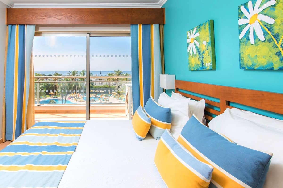 A hotel room with a bed, pillows, a view of a pool and resort, and artwork on the wall.