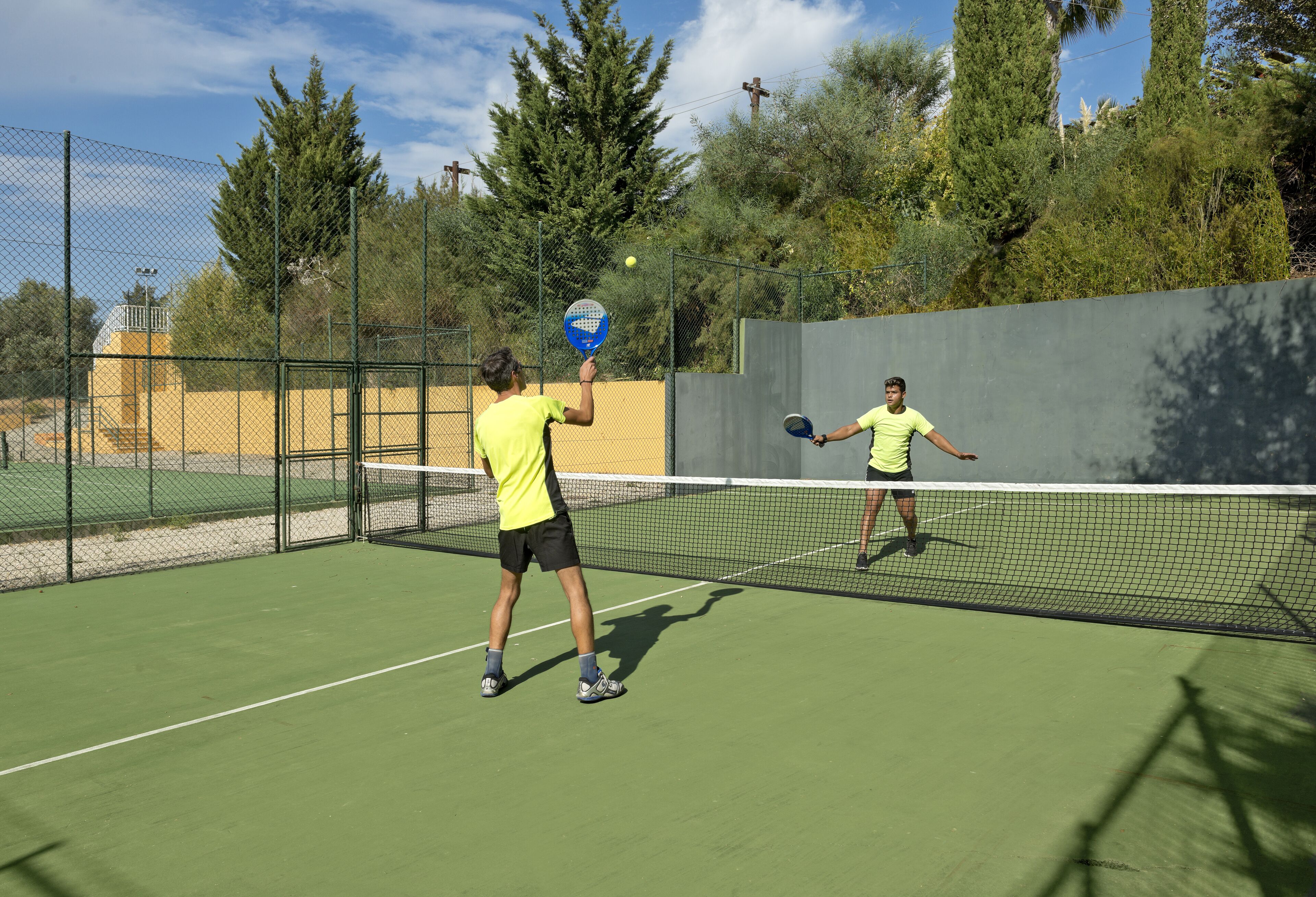 A tennis court with two players, a net, and surrounding fencing.