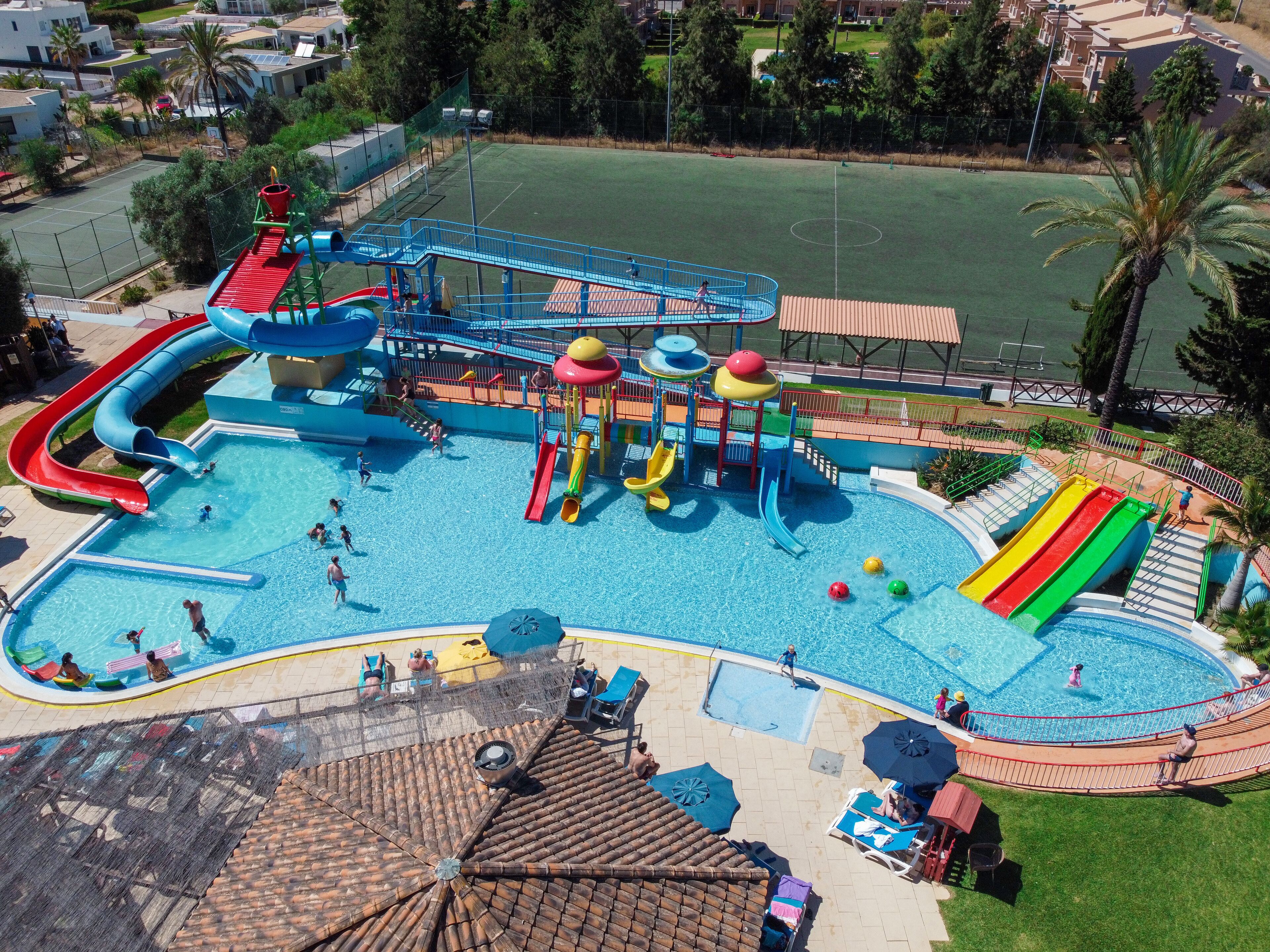 Een waterpark met meerdere glijbanen en zwembaden, omgeven door een omheind gebied, met een tennisbaan zichtbaar op de achtergrond.