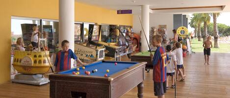 A pool table in a room with wooden flooring, surrounded by children playing.