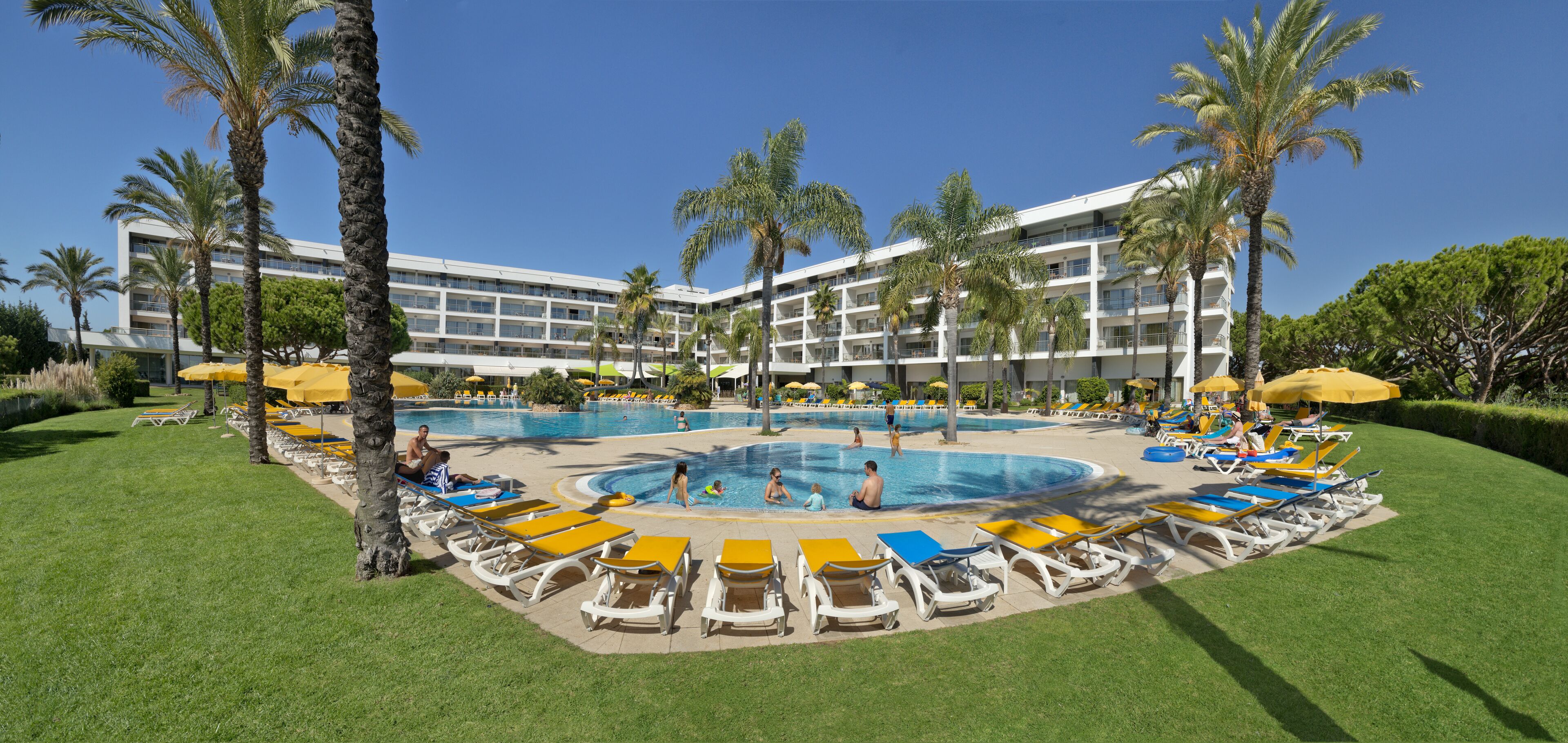 Ein Hotelpoolbereich mit mehreren Liegestühlen und Sonnenschirmen, umgeben von Palmen und einem großen Gebäude im Hintergrund.