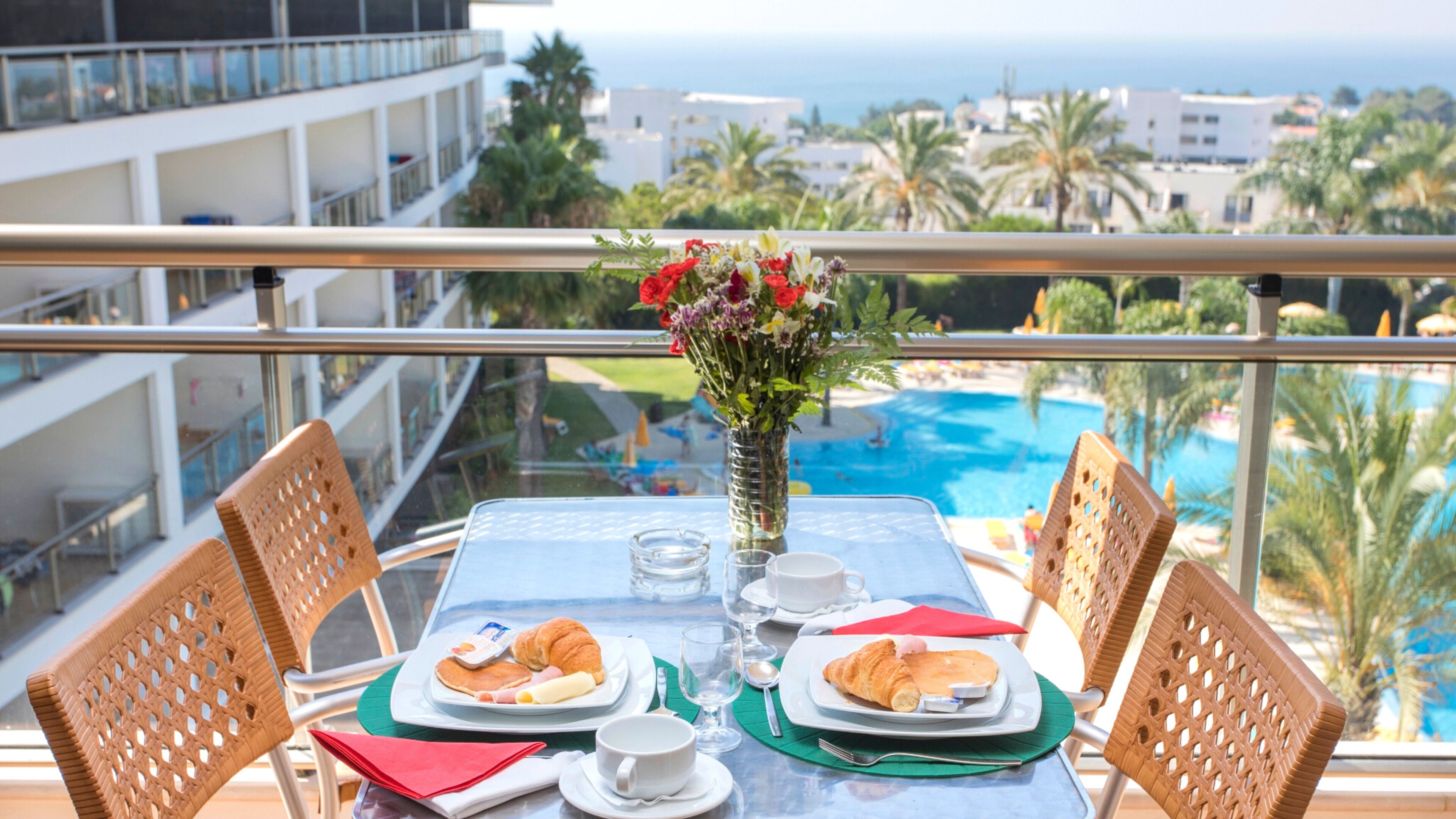 A dining table with breakfast items, a vase of flowers, and a view of a pool and buildings.
