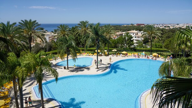 Ein großer, klarer, blauer Swimmingpool, umgeben von Palmen und Liegestühlen, mit Blick auf das Meer und Gebäude im Hintergrund.