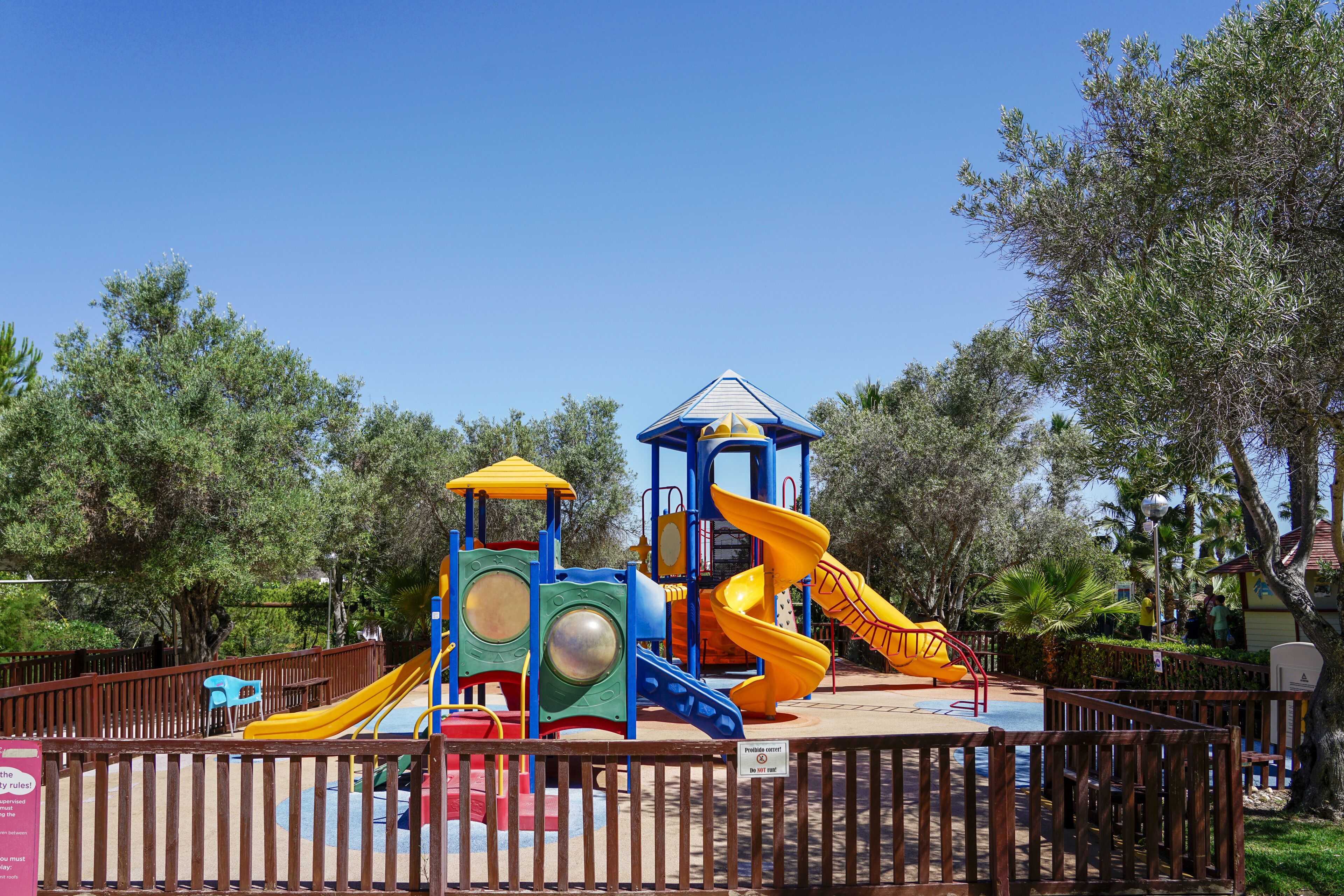 A playground with slides and climbing structures, surrounded by a wooden fence and trees.