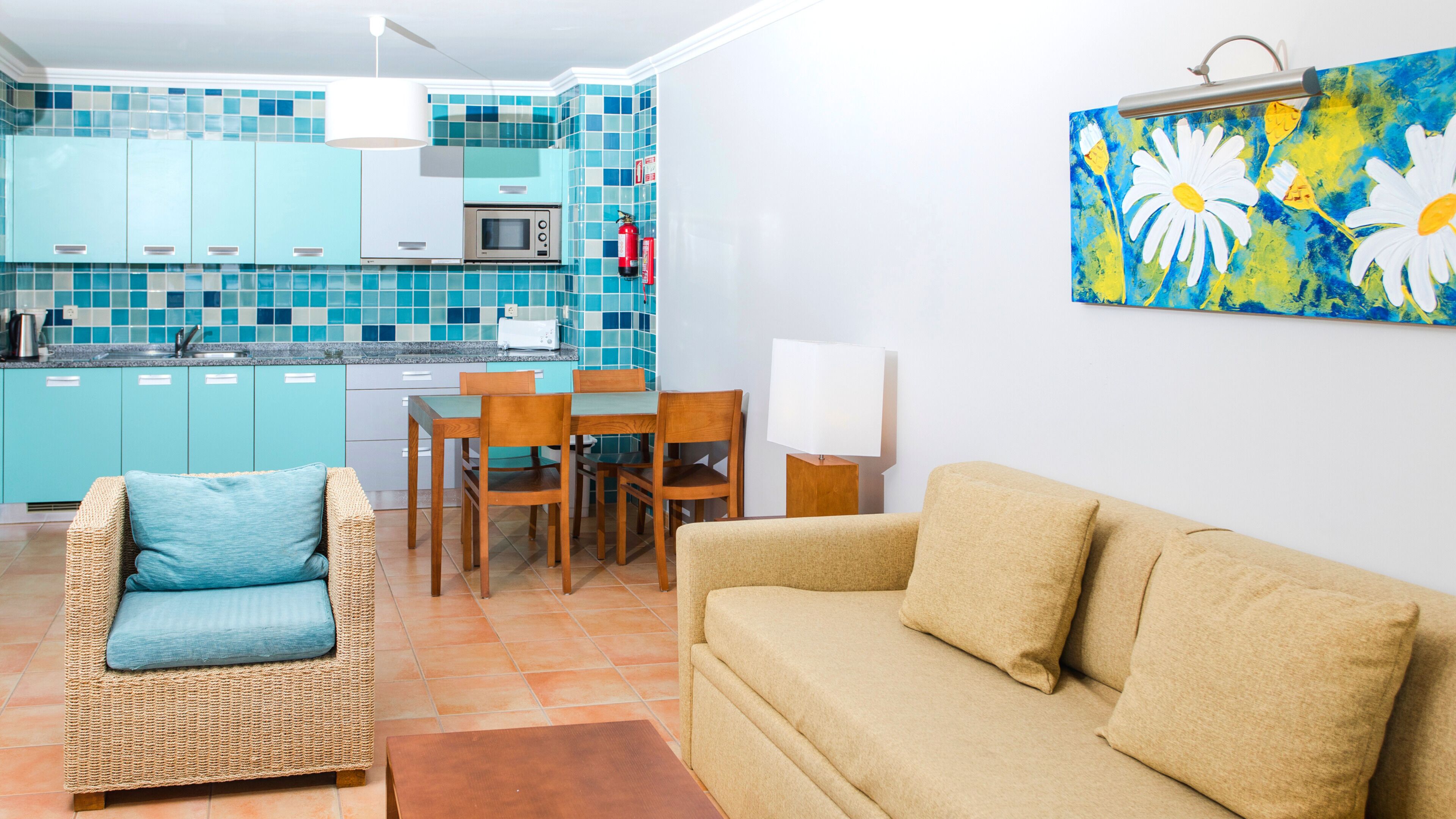 A living room with a beige sofa, a wicker chair with a blue cushion, a dining table with chairs, and a kitchen area with teal cabinets and a microwave.
