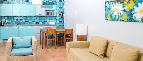 A living room with a beige sofa, a wicker chair with a blue cushion, a dining table with chairs, and a kitchen area with teal cabinets and a microwave.