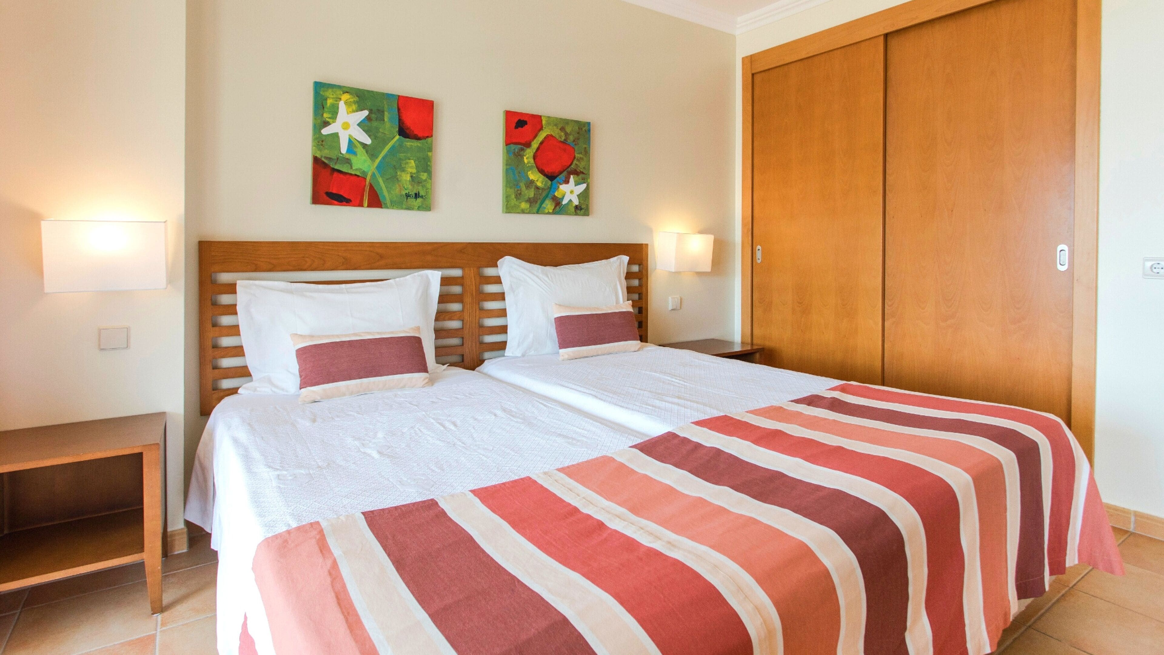 A double bed with a striped bedspread, two wall-mounted paintings, a wooden headboard, and a wooden wardrobe.