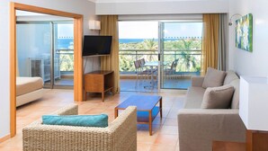 Ein geräumiger Wohnbereich mit einer Couch, einem Sessel, einem Fernseher und einem Balkon mit Meerblick.