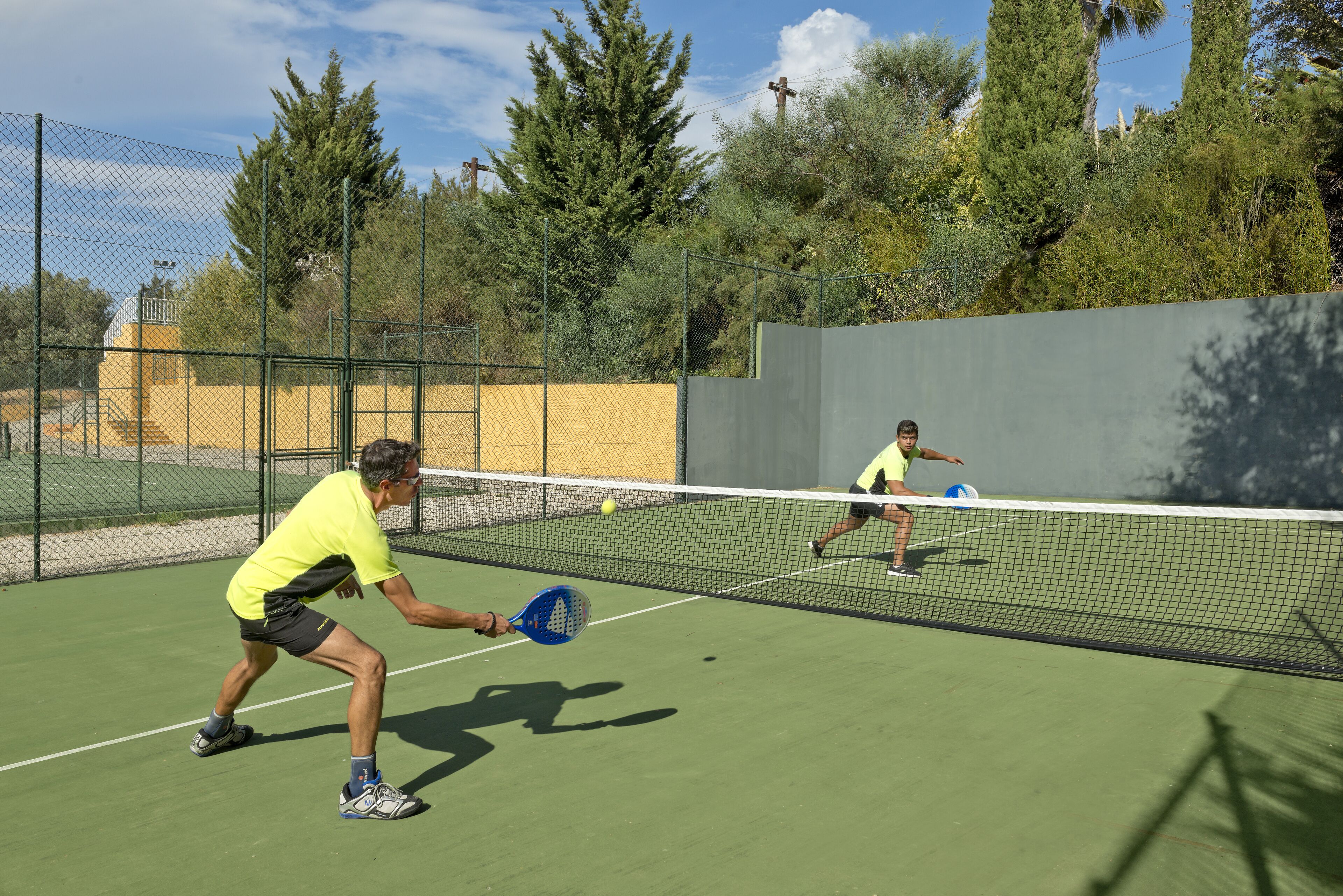 Een tennisbaan met net, omringd door een hek en bomen.