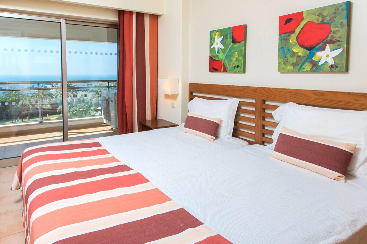 A hotel room with a large bed, a view of the ocean through a sliding glass door, and two abstract paintings on the wall.