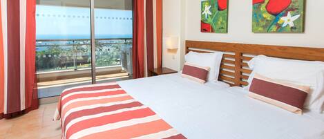 A hotel room with a large bed, a view of the ocean through a sliding glass door, and two abstract paintings on the wall.