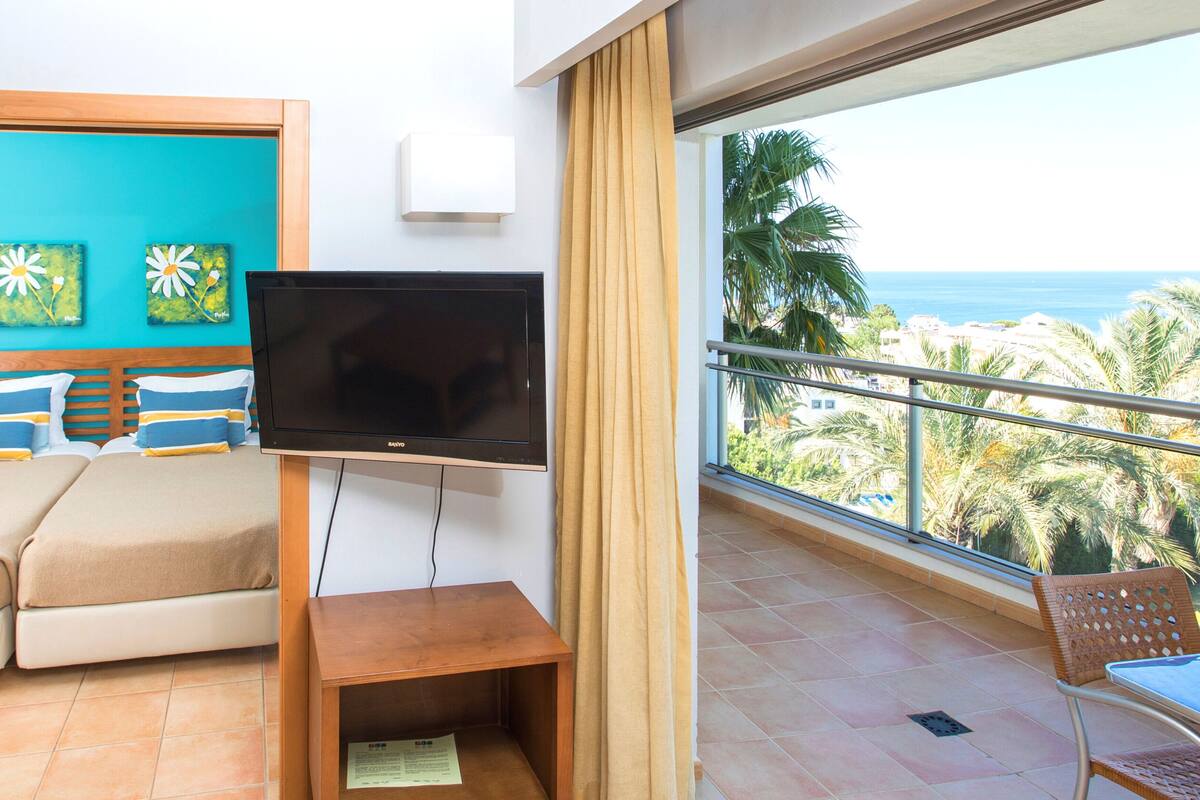 A hotel room with a bed, a TV, a balcony with a view of palm trees and the sea.