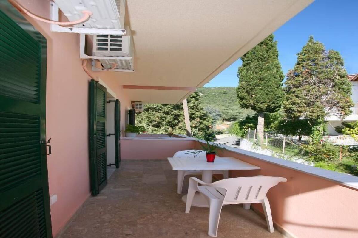 A balcony with a table and chairs, an air conditioning unit, and a view of trees and buildings.