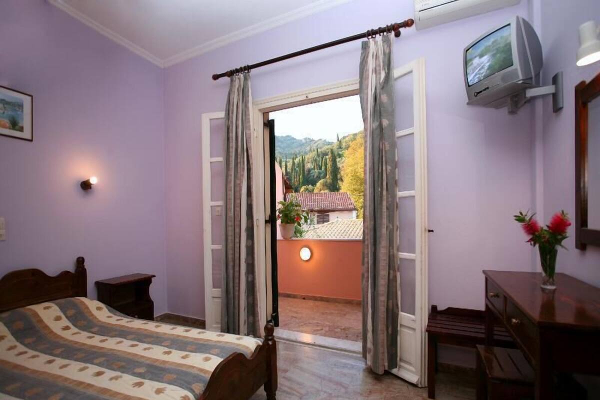A bedroom with a bed, a desk, a TV, and a view of a balcony with plants and a house.