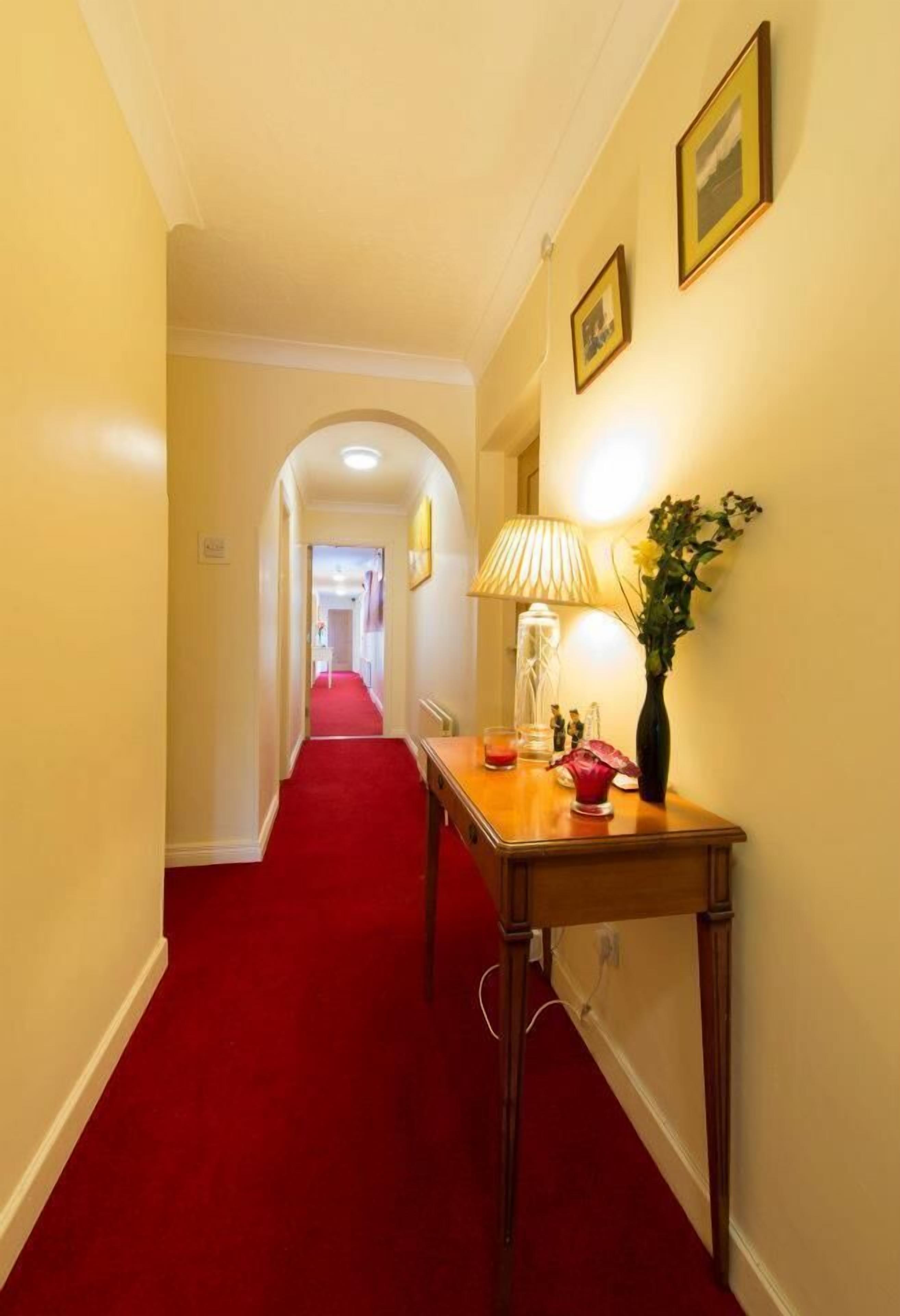 A hotel hallway with a red carpet, a wooden table with a lamp, and a vase with flowers.