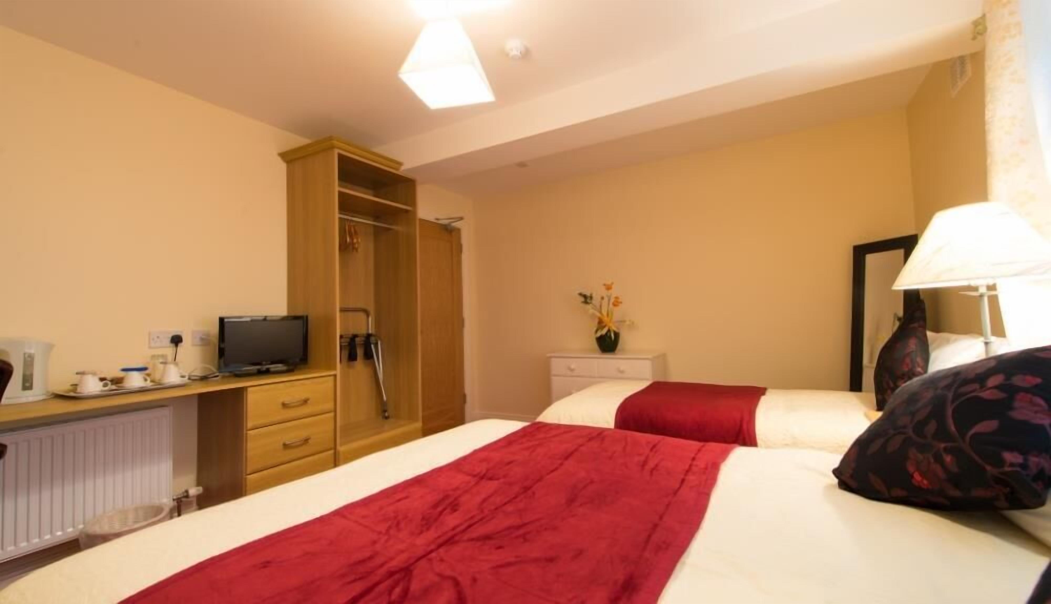 Quarto de hotel com cama, televisão em escrivaninha, guarda-roupa e mesa de cabeceira com abajur.