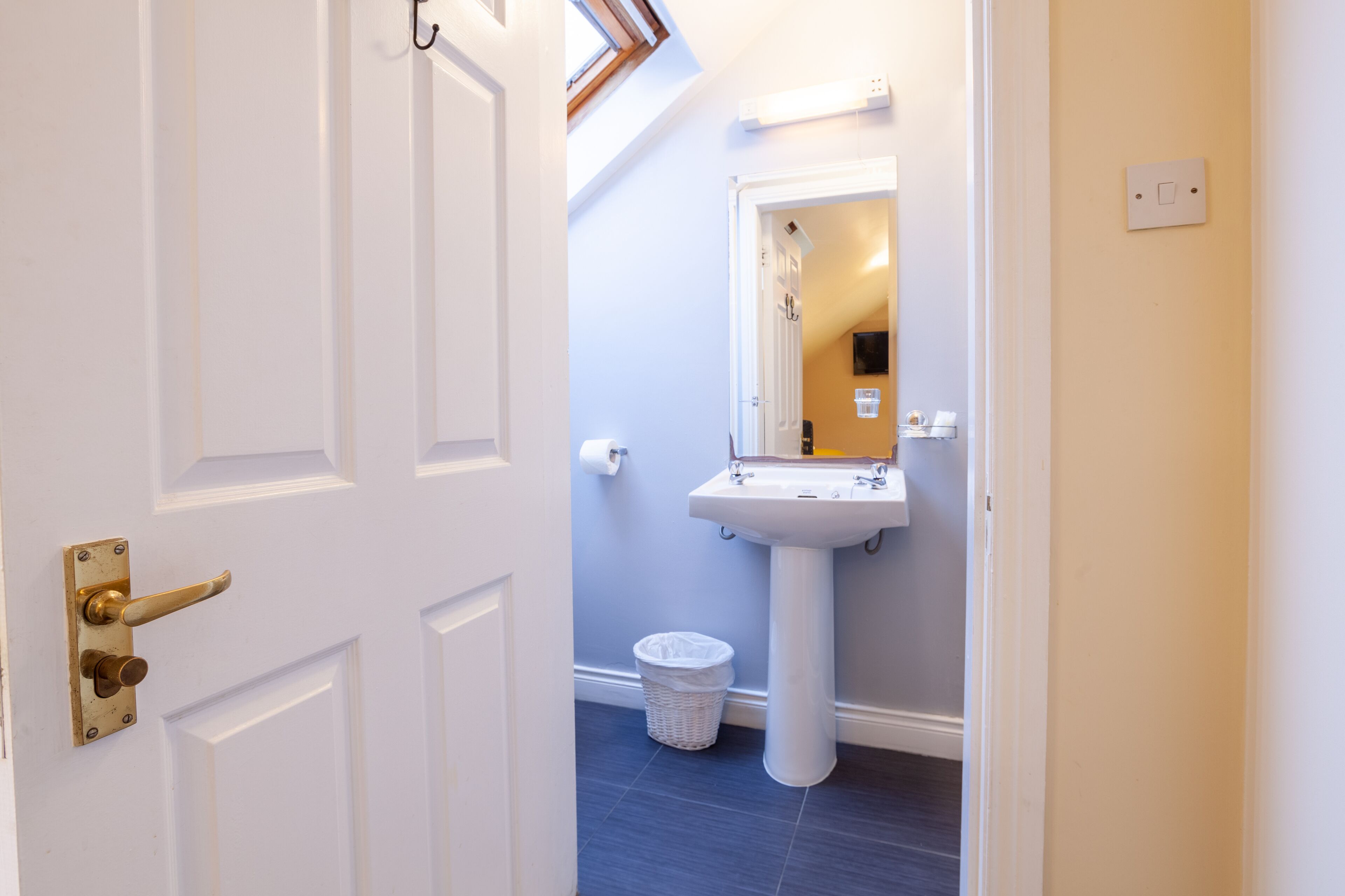 A bathroom with a white door, a sink, a mirror, and a trash bin.