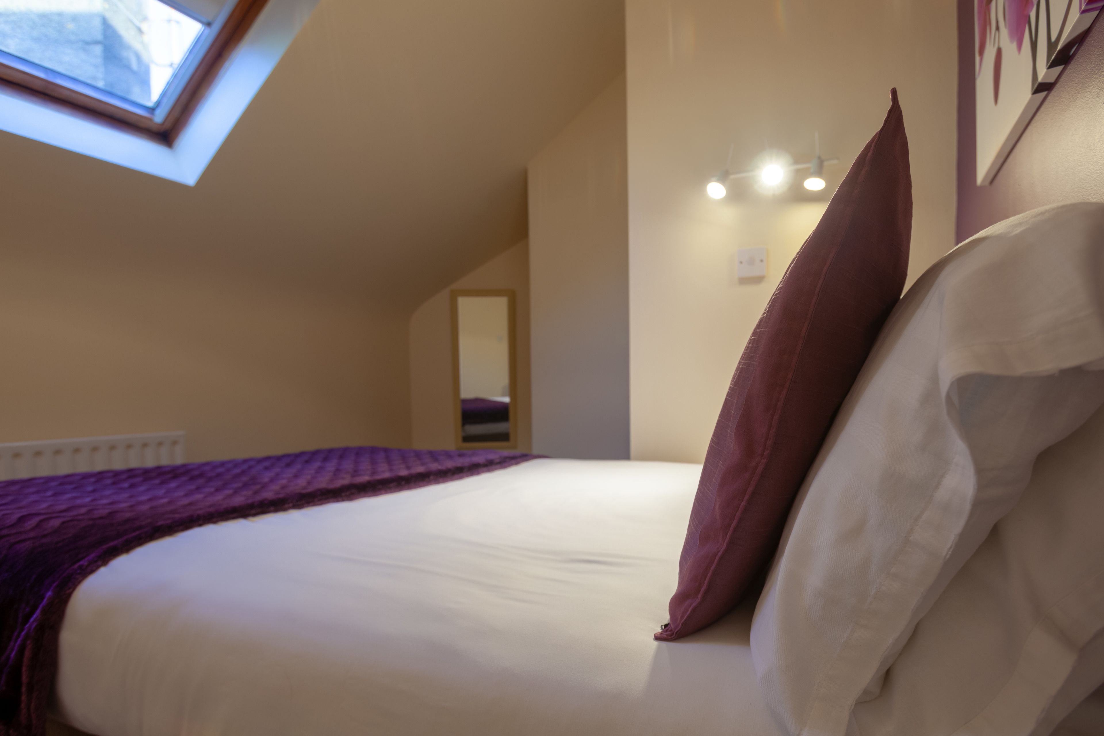 Un dormitorio con tragaluces, una cama con ropa de cama blanca y marrón, un edredón púrpura y una lámpara fijada a la pared.