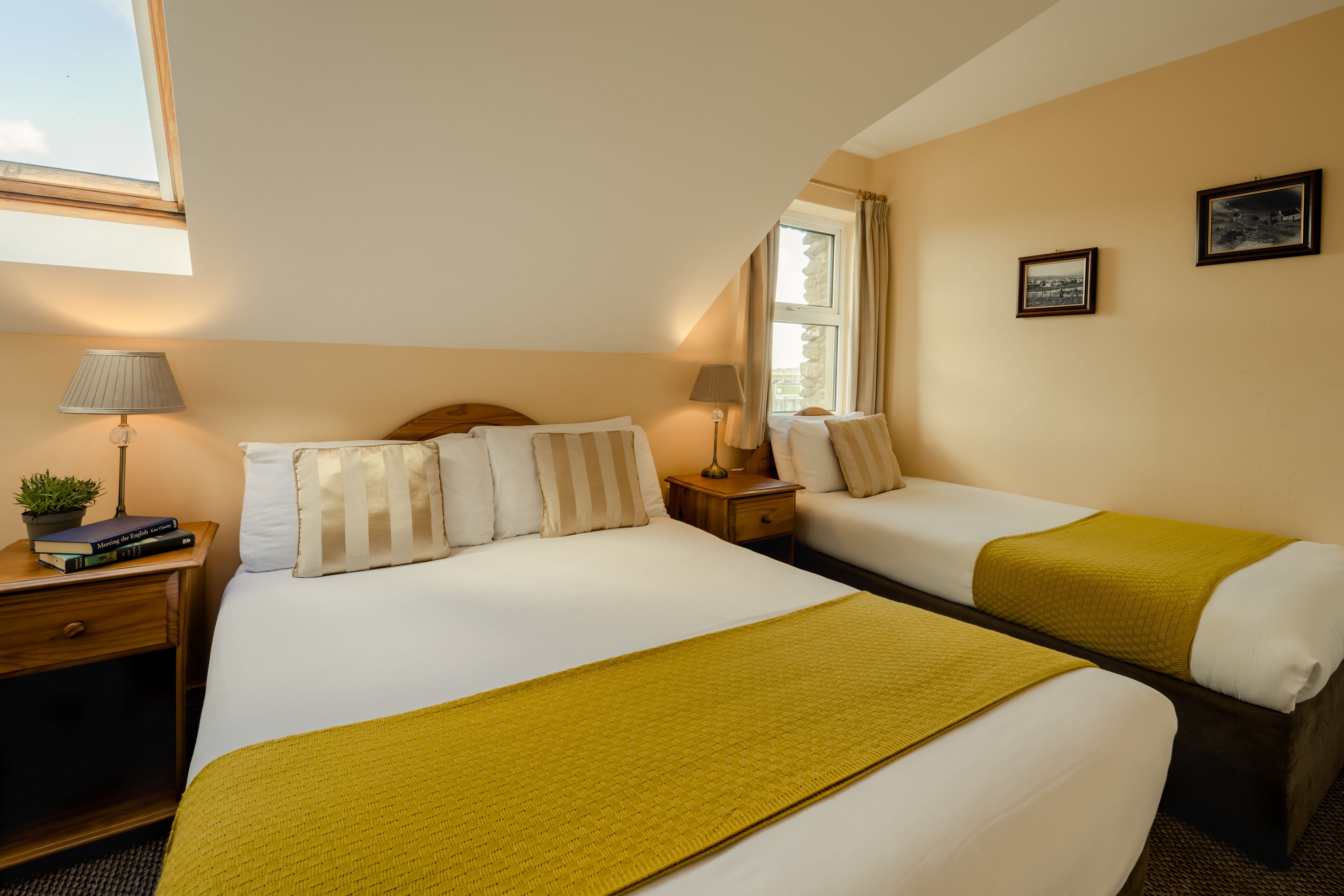 Habitación de hotel con una cama grande, una cama más pequeña, dos mesitas de noche con lámparas, una ventana con cortinas y un cuadro en la pared.