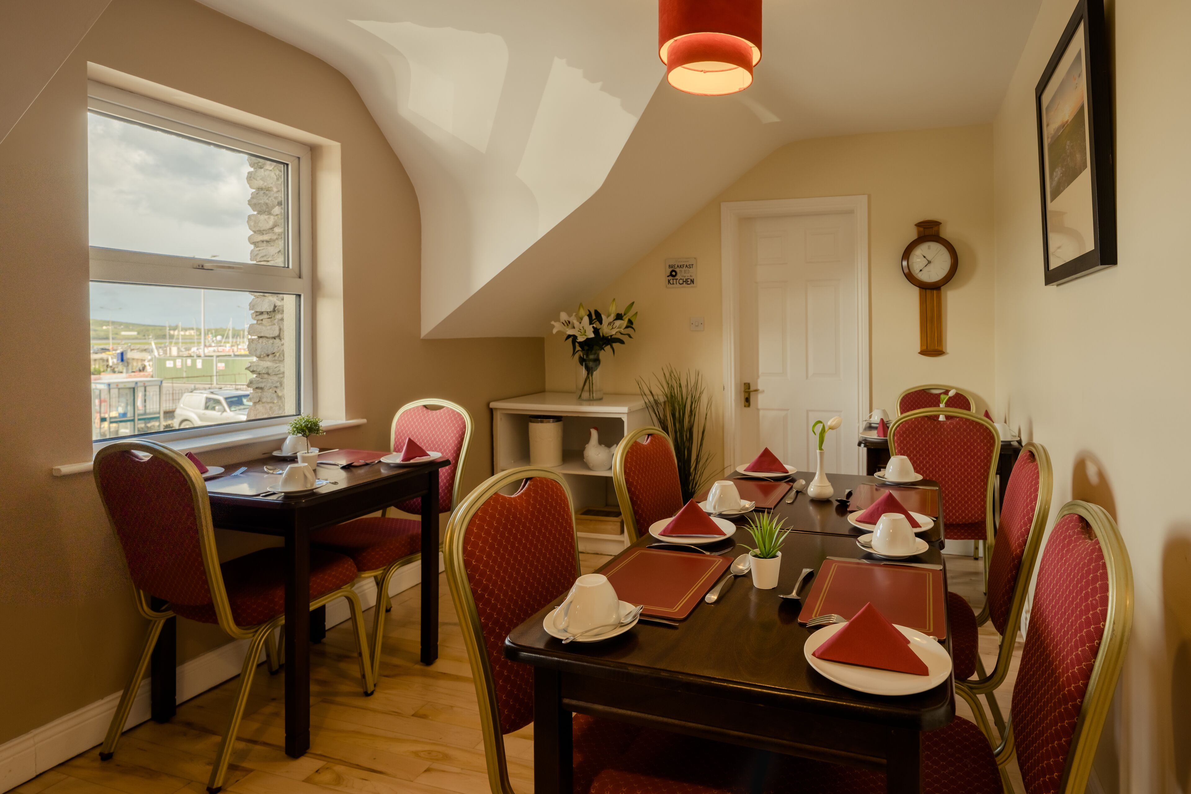 Un coin repas avec une table dressée pour six personnes, des chaises à revêtement rouge, un vase rempli de fleurs et une horloge au mur.