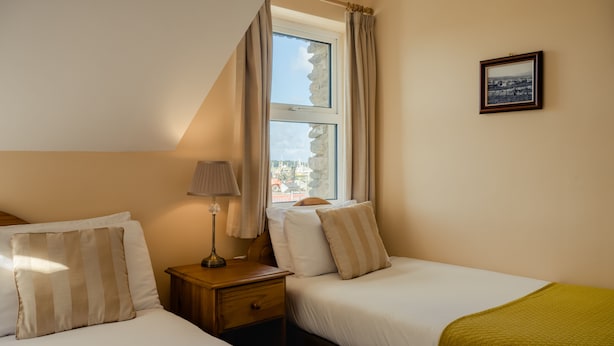 A hotel room with two beds, a nightstand, a lamp, a window with curtains, and a framed picture on the wall. - Quayside B&B (Dingle)