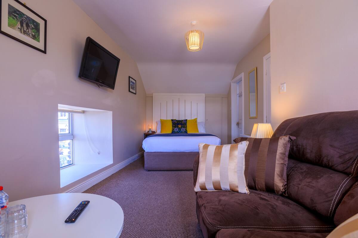 Una habitación de hotel con una cama, un sofá de cuero marrón, una mesa blanca, un televisor y una ventana.
