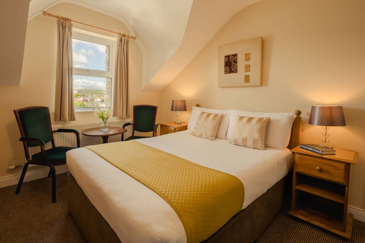 Una habitación de hotel con una cama grande, dos sillas, una mesita y un ventanal con cortinas.