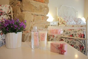 Un dormitorio con pared de piedra, ropa de cama floral, estructura de cama de metal blanco y una mesita de noche con un jarrón de flores moradas, dos botellas de productos 'Romantic Home' y una caja de regalo rosa.