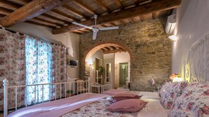 A bedroom with a stone wall, wooden beams, a ceiling fan, and floral bedding. - Il Vignolino Bed and Breakfast (Barberino di Mugello)