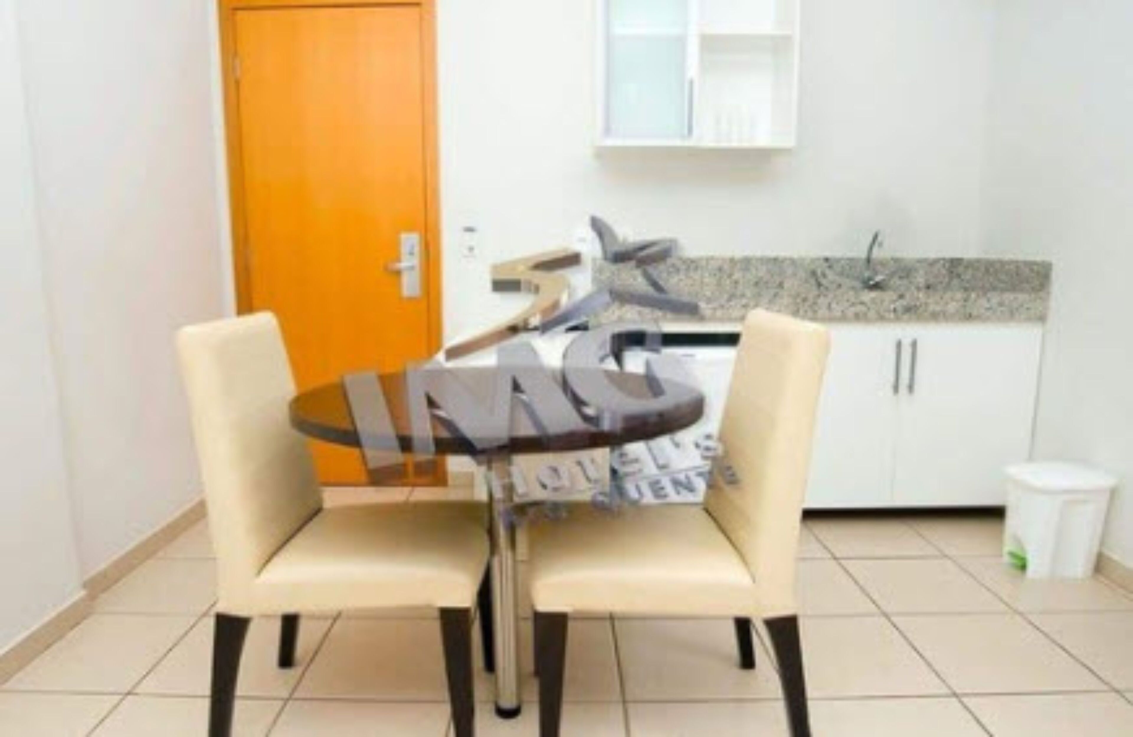 Un pequeño comedor con dos sillas y una mesa, una cocina al fondo y un cubo de basura.