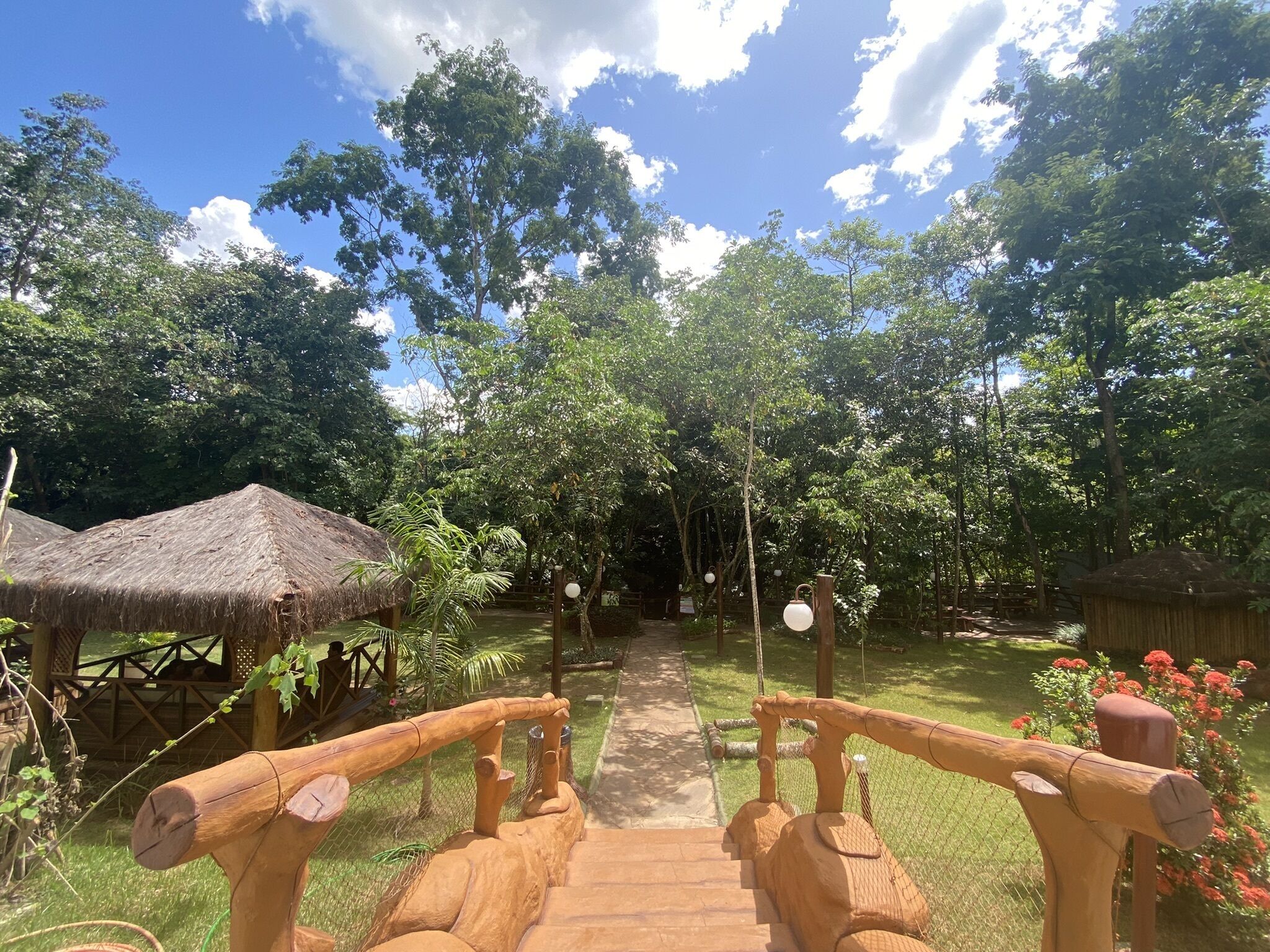 A wooden pathway leading through a lush garden with thatched-roof structures and a clear sky.