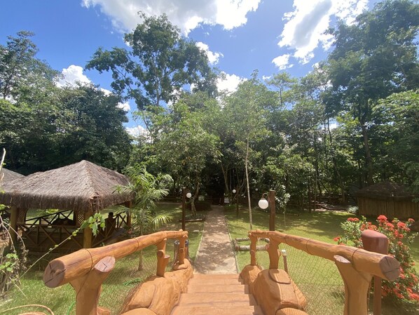 Un sendero de madera que serpentea por un exuberante jardín con construcciones de techo de paja y un cielo despejado.