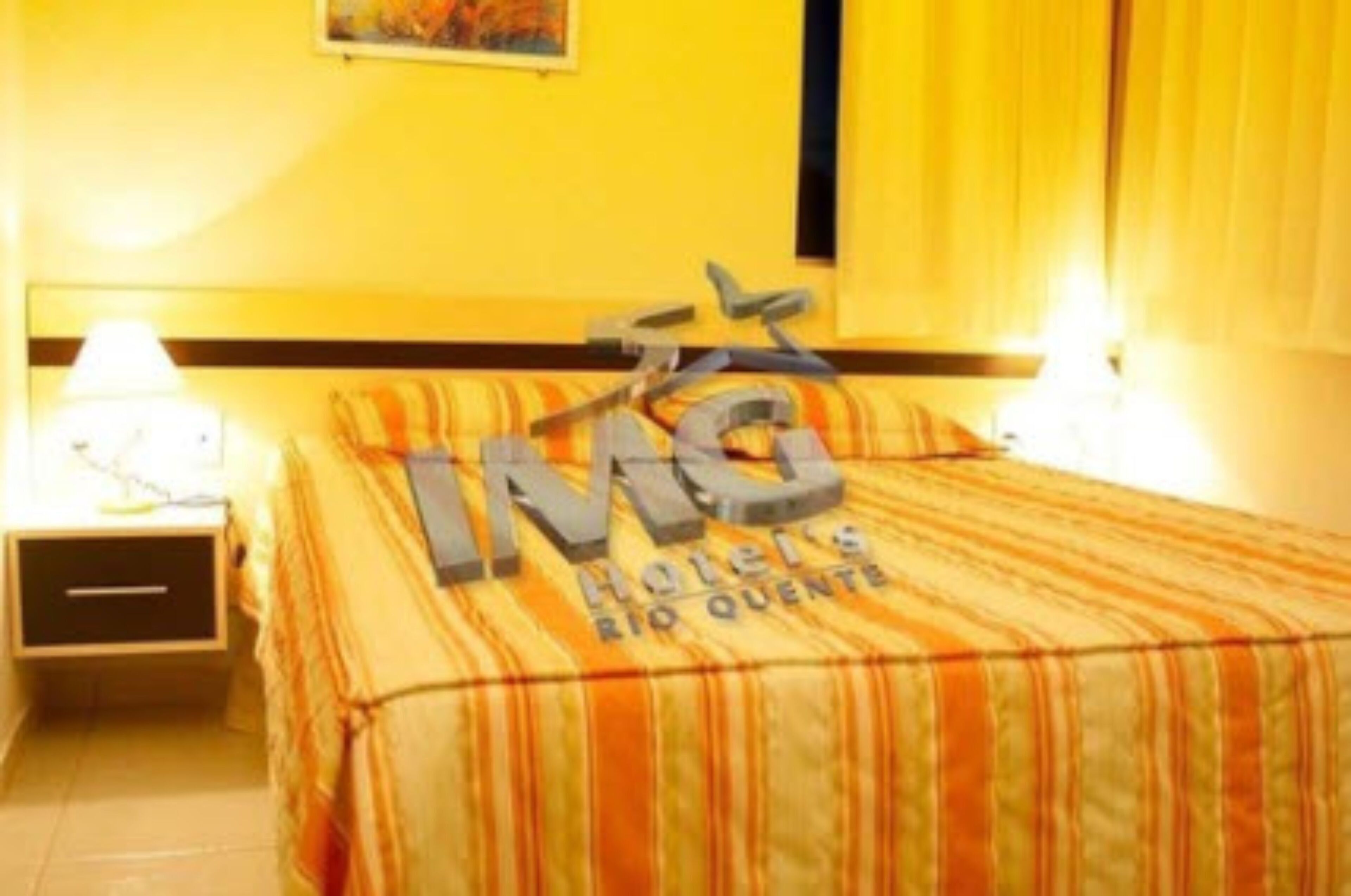 A hotel room with a bed, a nightstand, and a lamp. The room has a striped bedspread and a sign that reads "IMG Hotels Rio Quente".