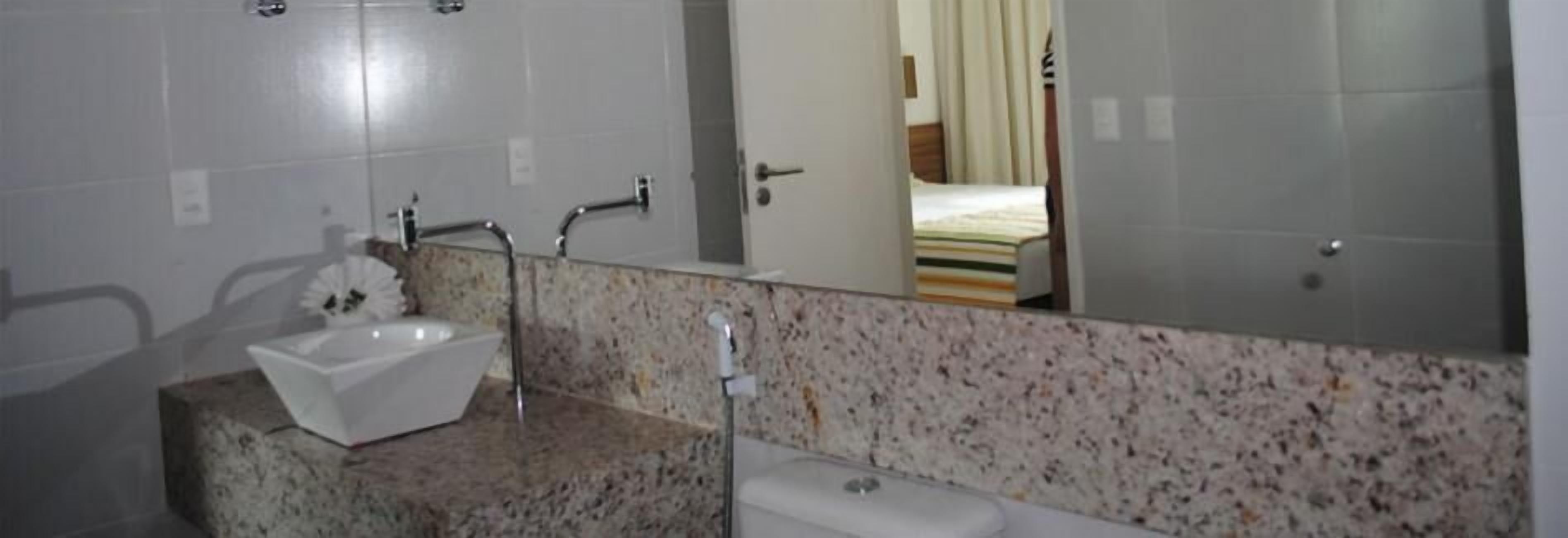 Casa de banho com lavatório branco, bancada de granito e espelho a refletir uma cama.