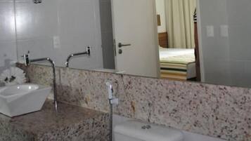 Casa de banho com lavatório branco, bancada de granito e espelho a refletir uma cama.