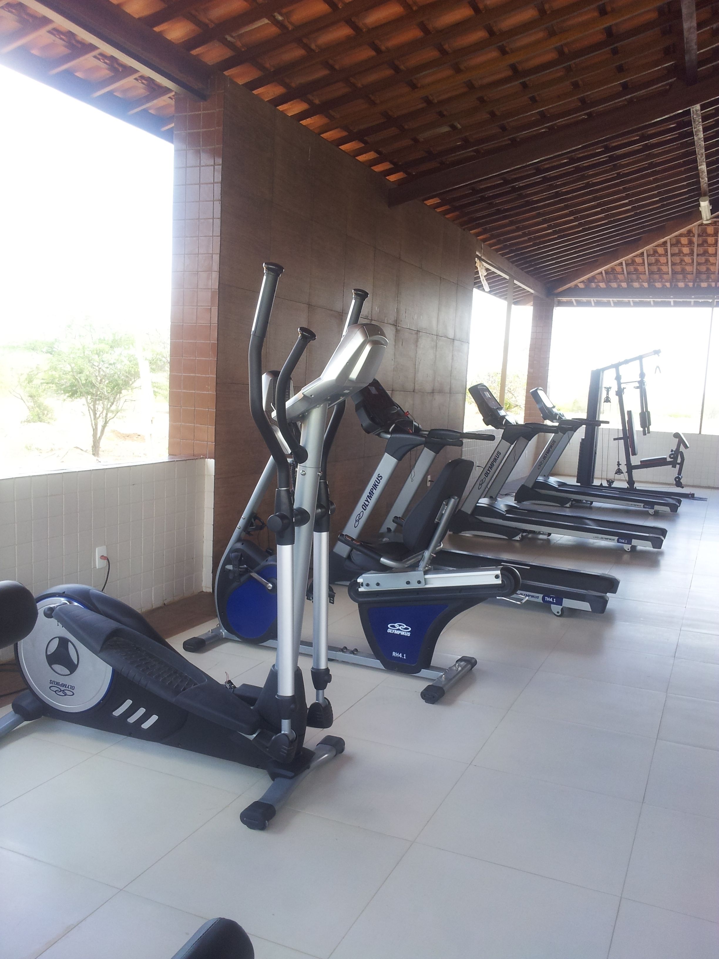 A gym with multiple exercise machines, including treadmills and elliptical trainers, under a covered area with a tiled floor and brick walls.