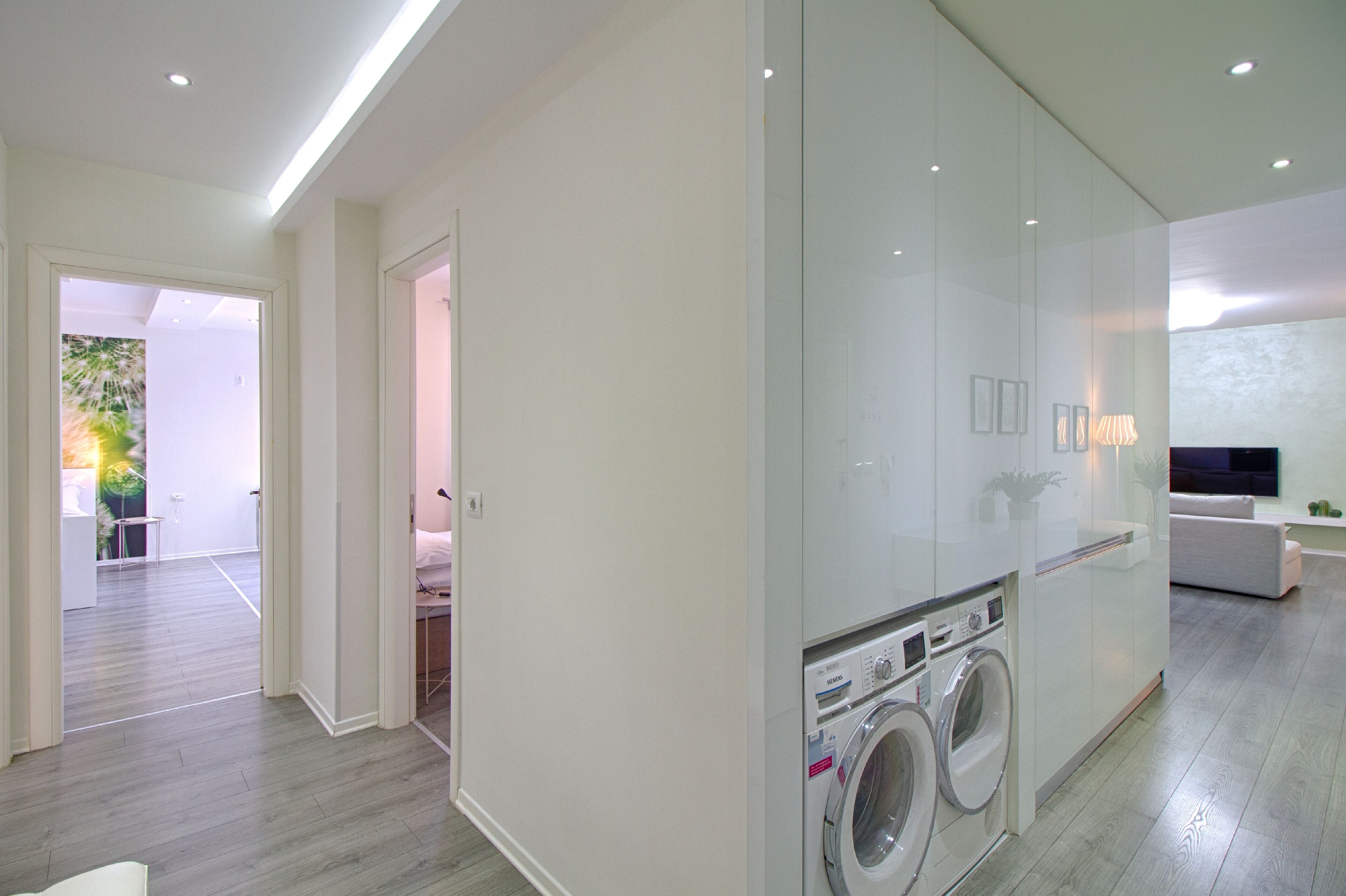 A modern laundry room with a washing machine and dryer, a glass partition, and a view into a living area with a sofa and a TV.