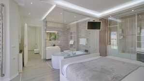 A modern bedroom with a large bed, a glass-enclosed shower, a vanity with a mirror, and a television mounted on the wall.
