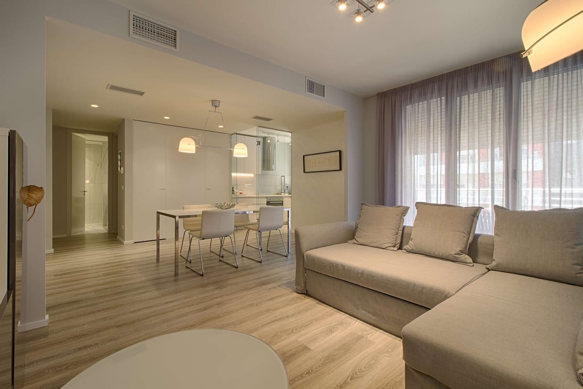 Una sala de estar moderna con un sofá color beige, un comedor con una mesa y sillas, y una cocina al fondo.