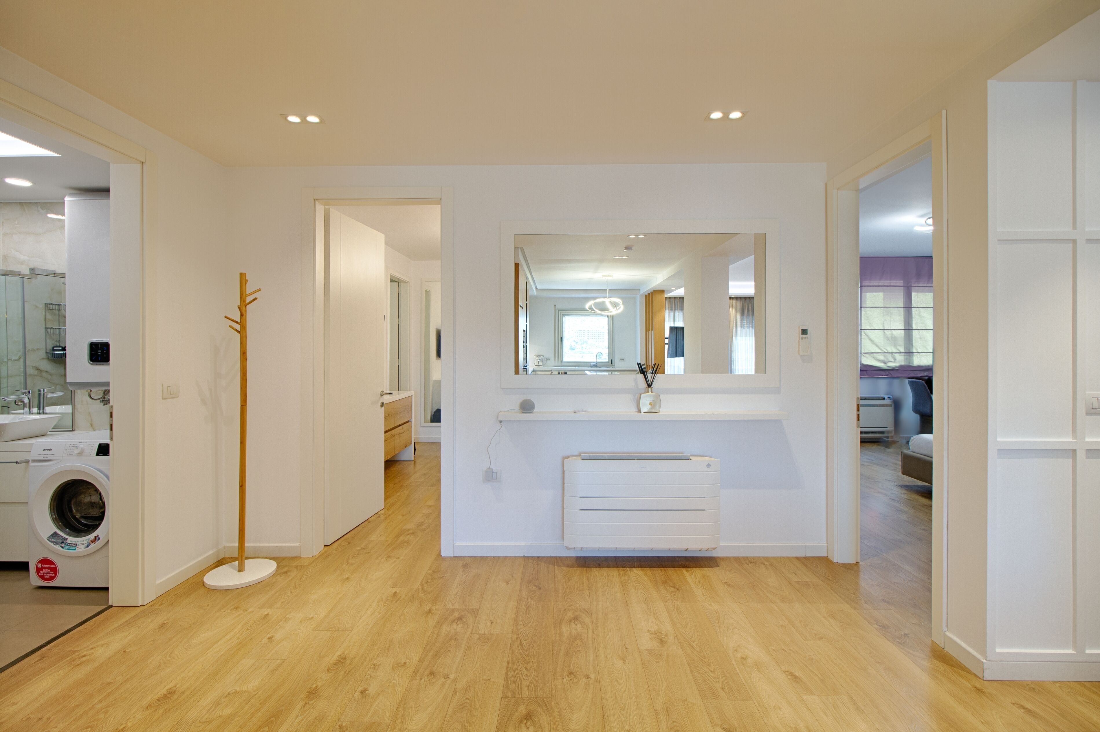 A modern, well-lit room with a wooden floor, a built-in washing machine, a standing heater, and a large mirror reflecting the room.