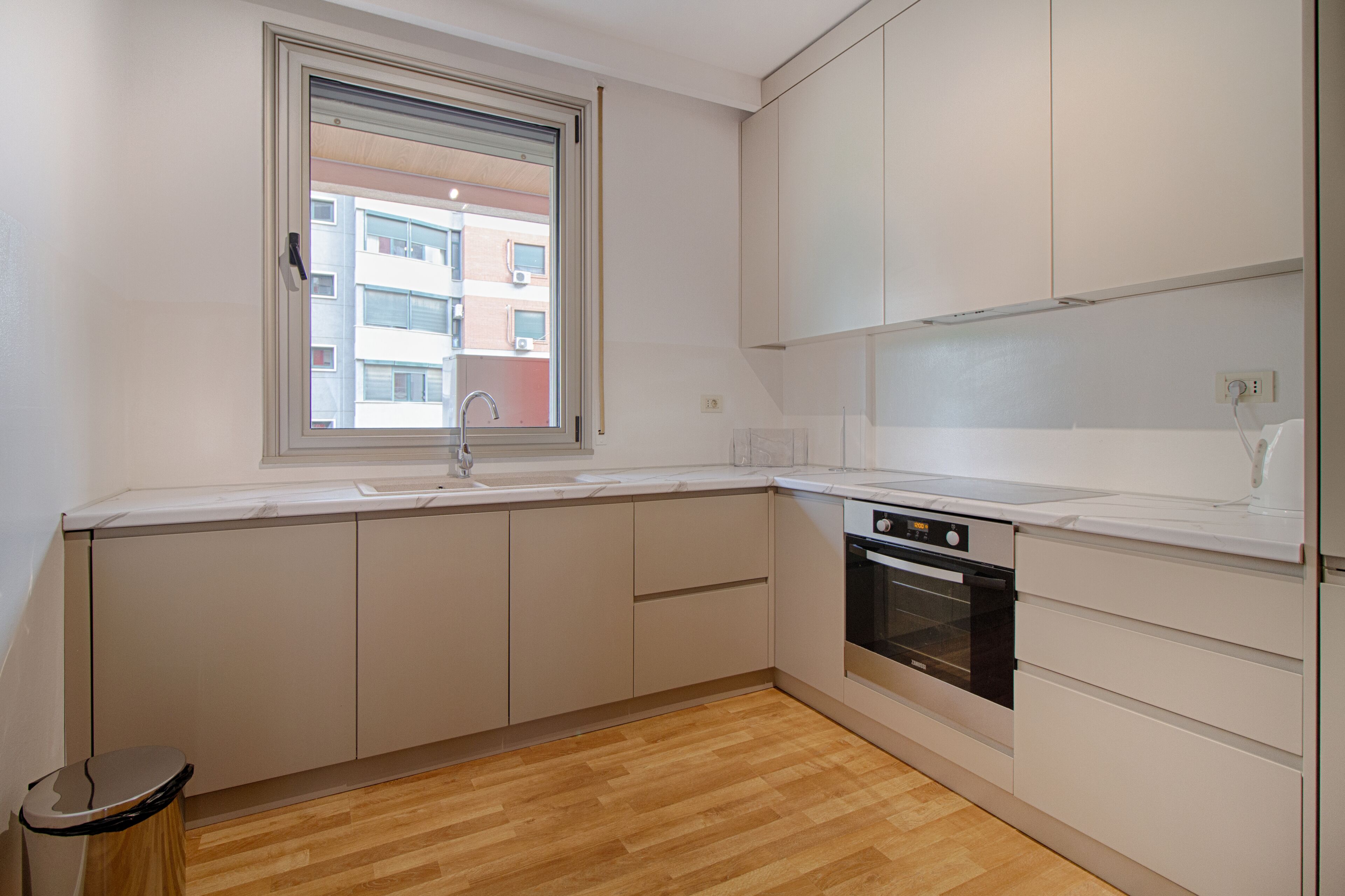 Une cuisine moderne avec des armoires blanches, un four encastré et une fenêtre donnant sur un bâtiment à l’extérieur.