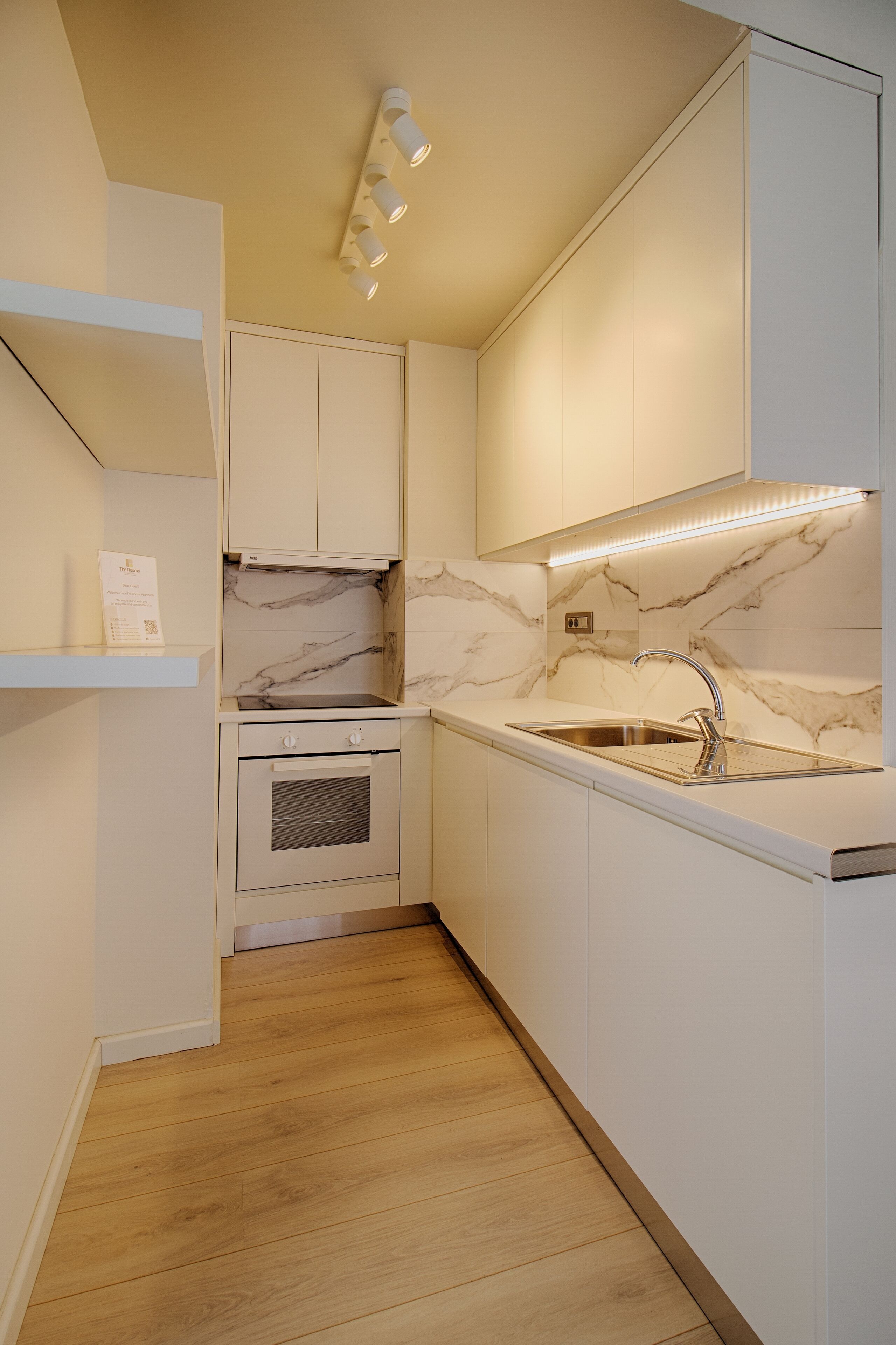 Una cocina compacta con armarios blancos, protector de encimera de mármol y pileta de acero inoxidable.