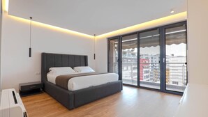 A modern bedroom with a large bed, a small bedside table, a built-in bench, and a sliding glass door leading to a balcony.