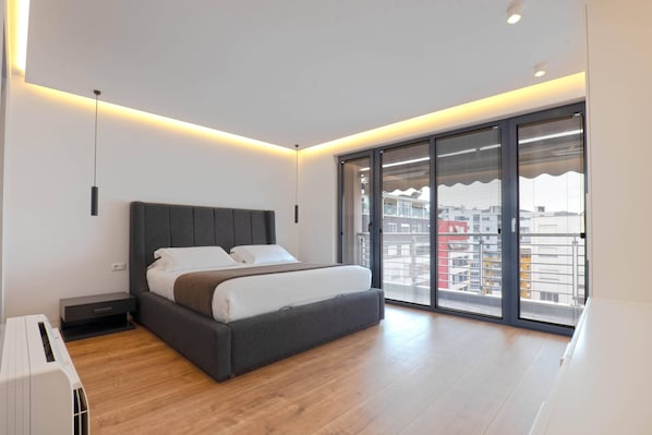 A modern bedroom with a large bed, a small bedside table, a built-in bench, and a sliding glass door leading to a balcony.