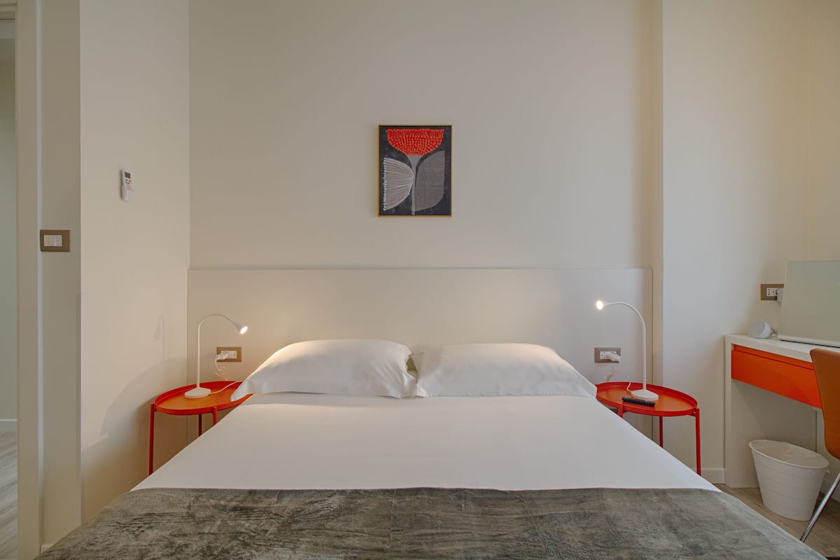 Una habitación de hotel moderna con una cama grande, dos mesitas de noche con lámparas, un escritorio con silla y una obra de arte colgada en la pared.