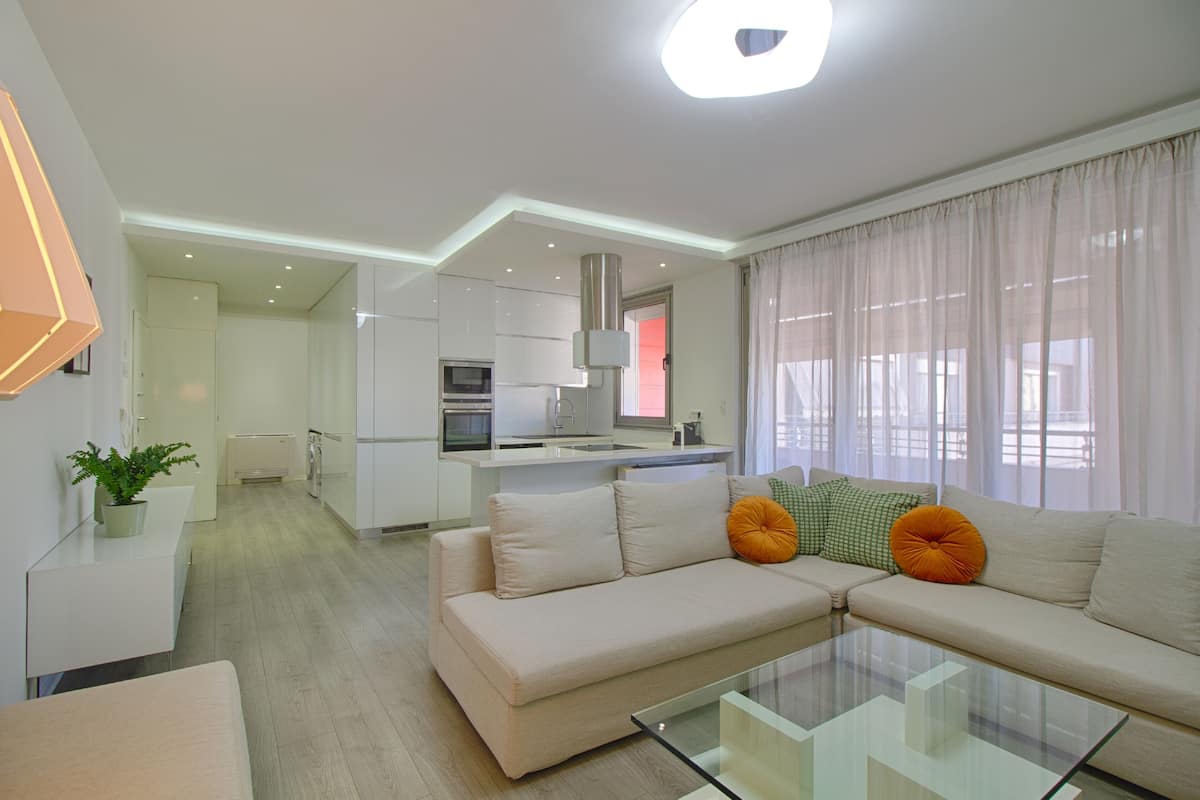 Una sala moderna con un sofá, una mesa de centro de cristal y una cocina al fondo.