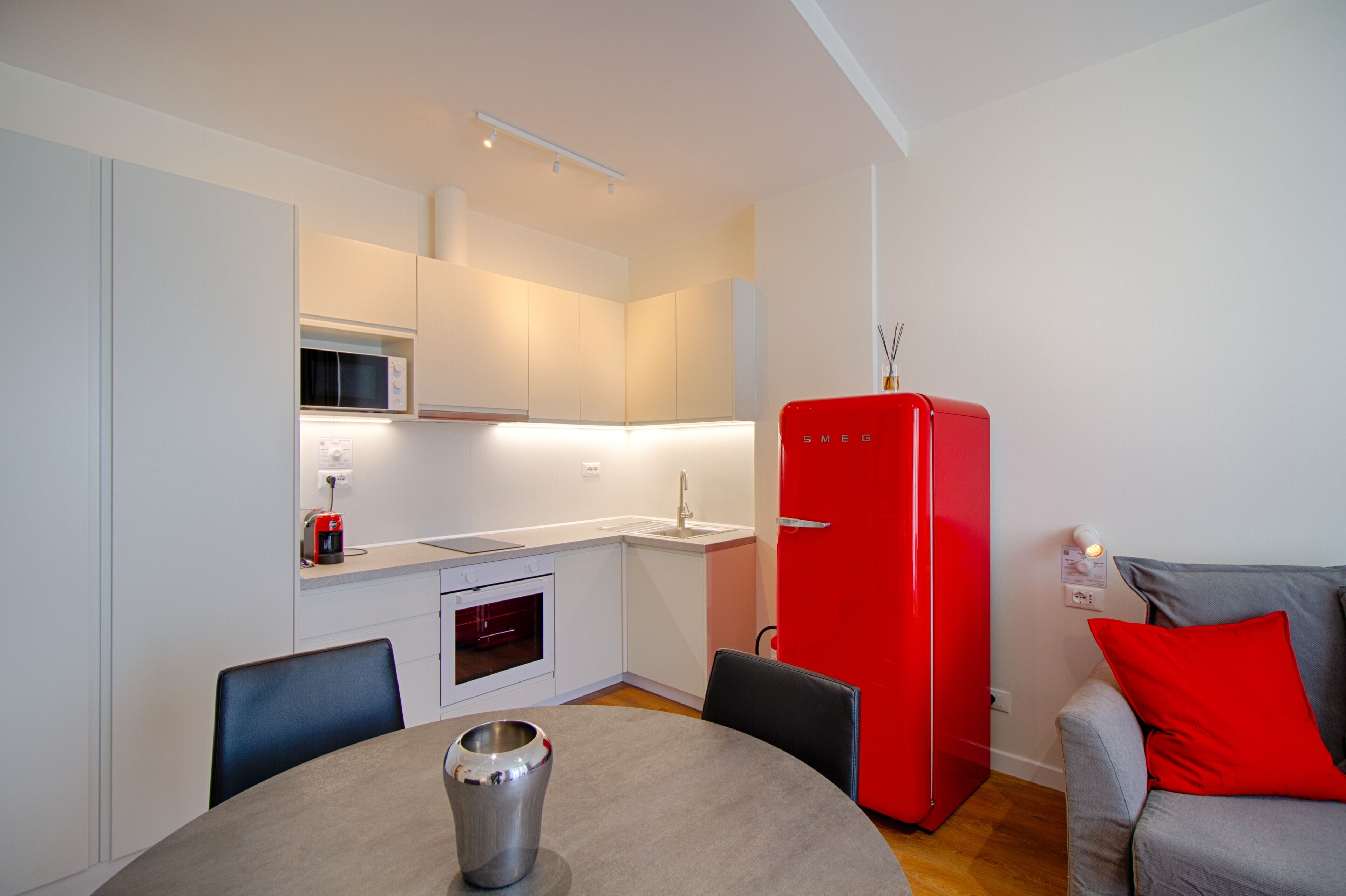 A compact kitchen with white cabinetry, a red refrigerator, a microwave, and a small oven. There is a dining table with chairs and a vase.