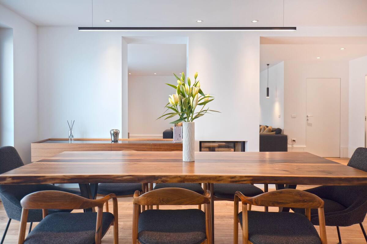 Un comedor moderno con una larga mesa de madera, sillas tapizadas oscuras y un florero con flores.