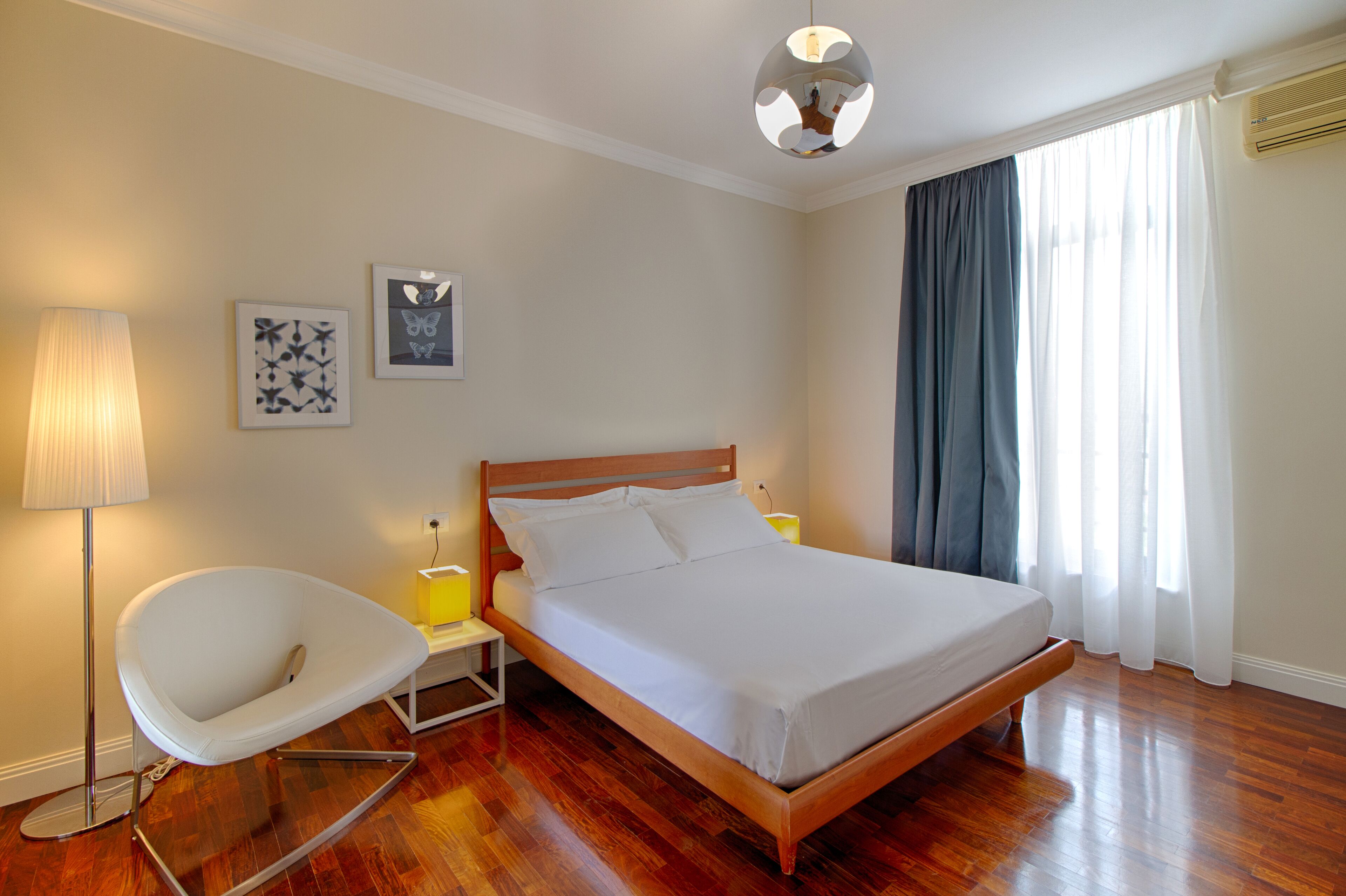 A bedroom with a wooden bed, a white chair, a floor lamp, and two wall art pieces.