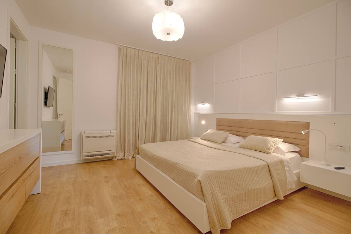 Un dormitorio con una cama grande, mesitas de noche, un espejo, una lámpara en el techo y una ventana con cortinas.